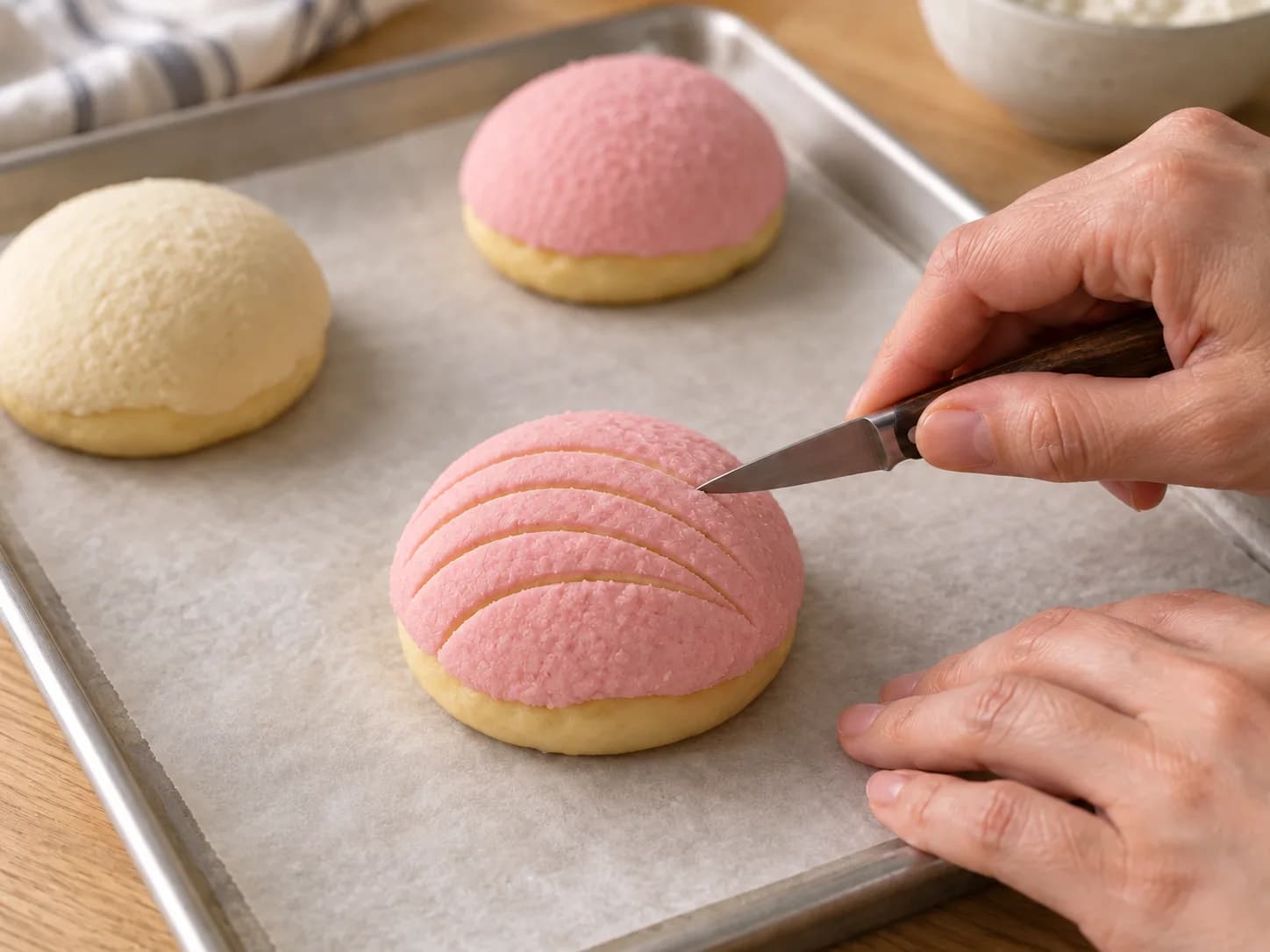 A knife tip scores curved lines in a pink topping cap, with pink and plain conchas behind it.