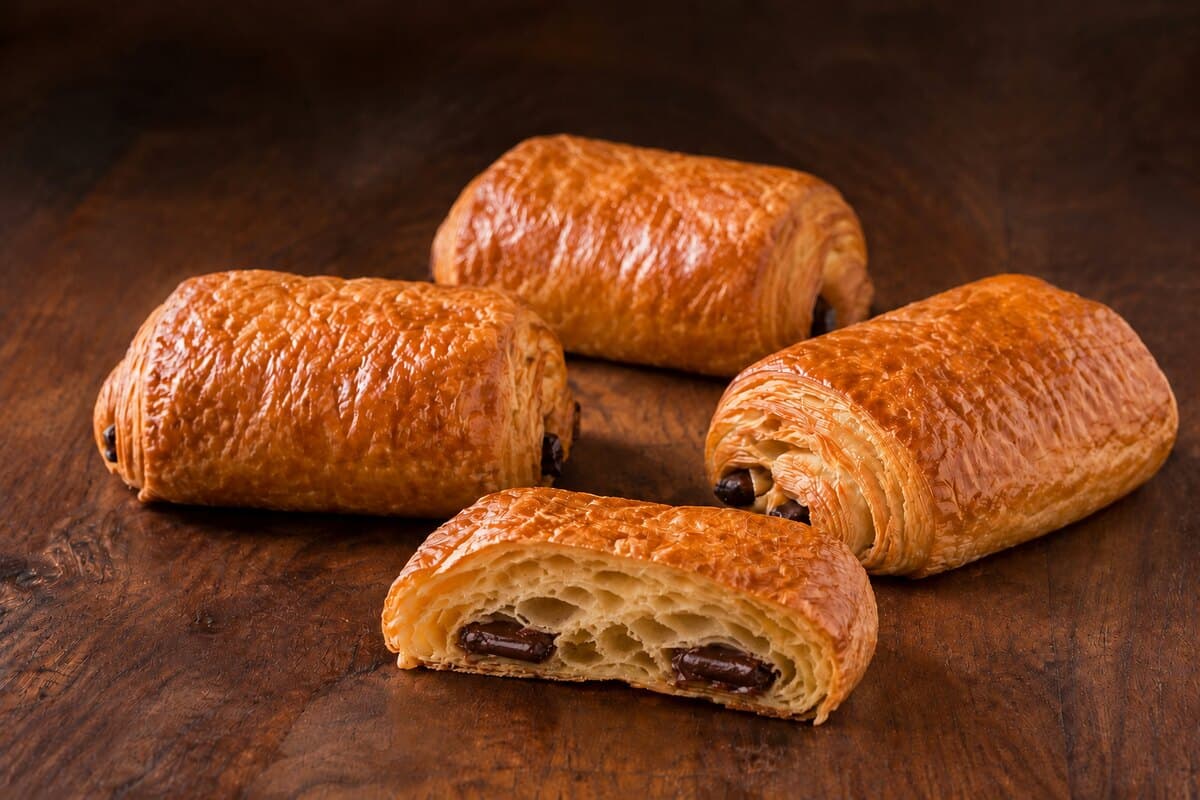 Four golden rectangular pains au chocolat made from laminated dough, one cut open to show the airy layers and two chocolate batons.